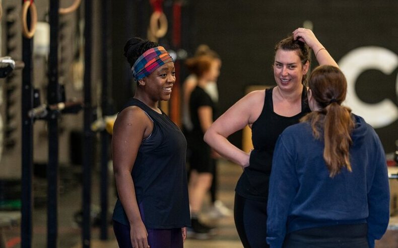 Three women chatting and smiling between sets at a CrossFit gym